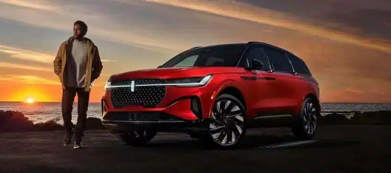 Man stands beside a red SUV at sunset on a coastal road, highlighting the car's sleek design.