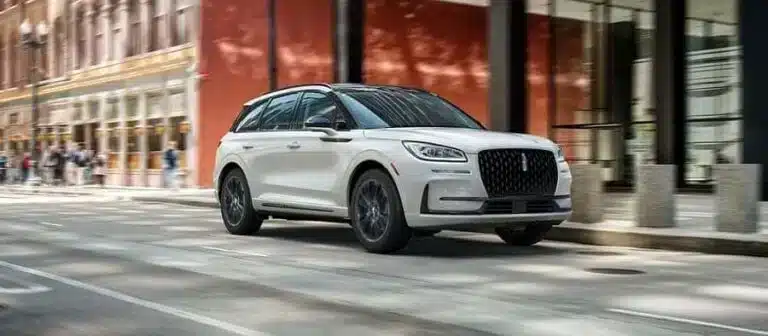 A white SUV driving on an urban street past historic brick buildings, showcasing its sleek design.