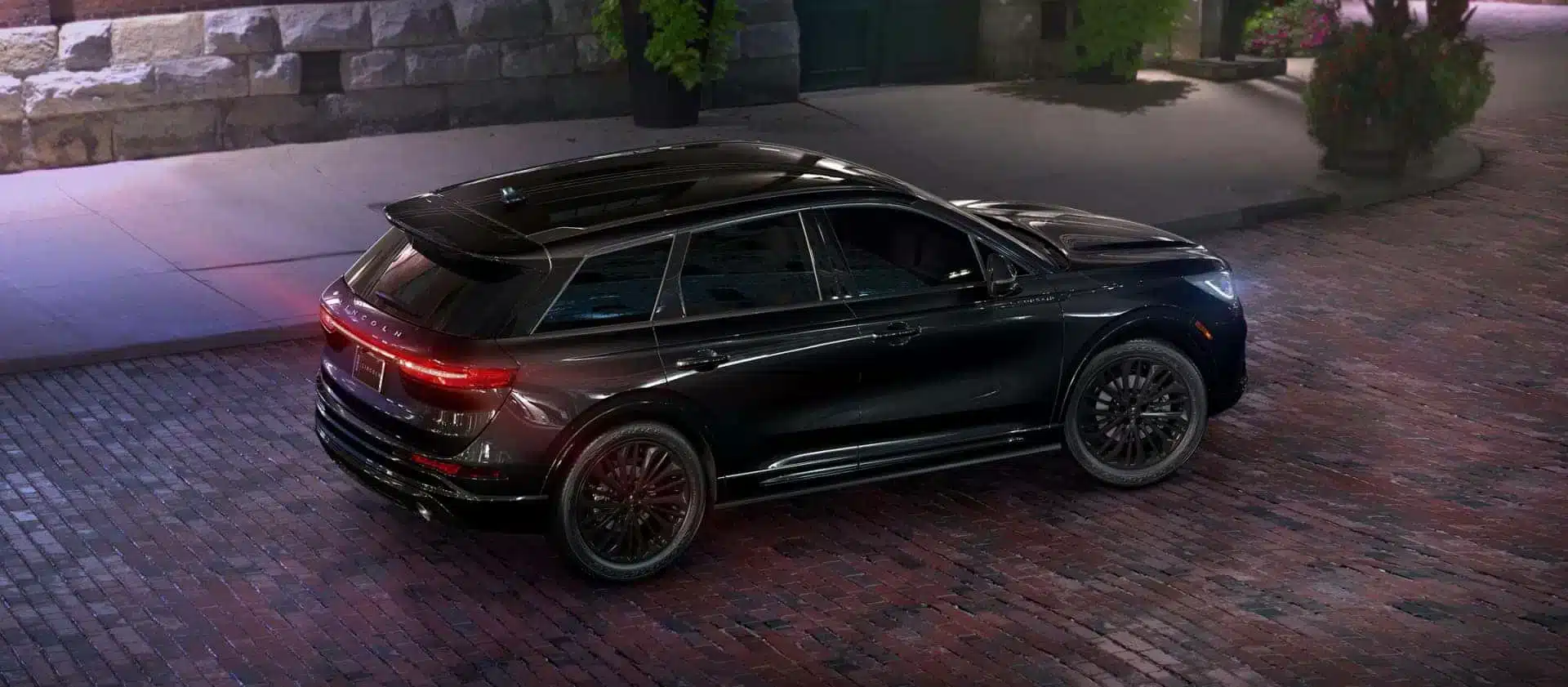 Black SUV parked on cobblestone street at night, rear-side view, illuminated by streetlights.