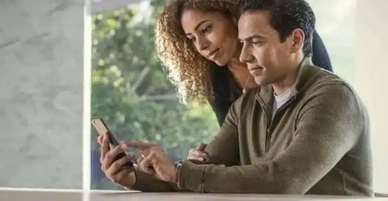 Couple using a smartphone together, sitting indoors with a green outdoor view.