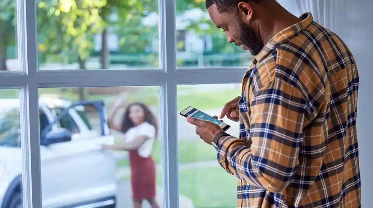 Man using smartphone near window; woman outside by a parked car. Bright, sunny day.