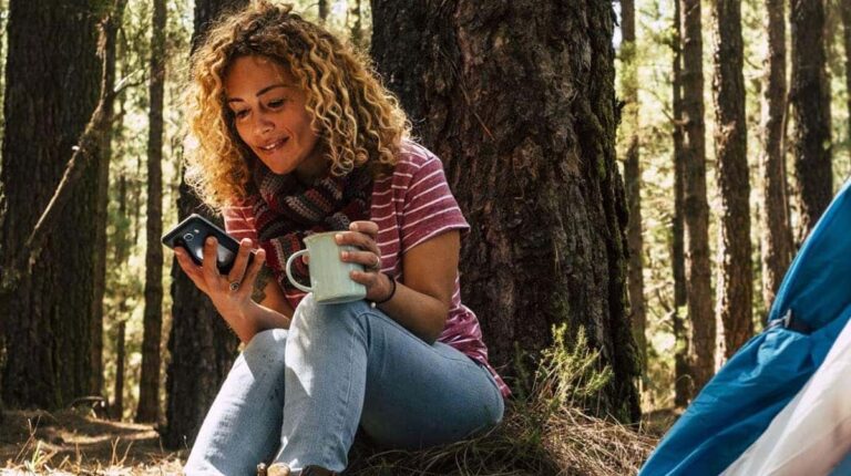 Woman with curly hair enjoys coffee in a forest, using her phone near a tent.