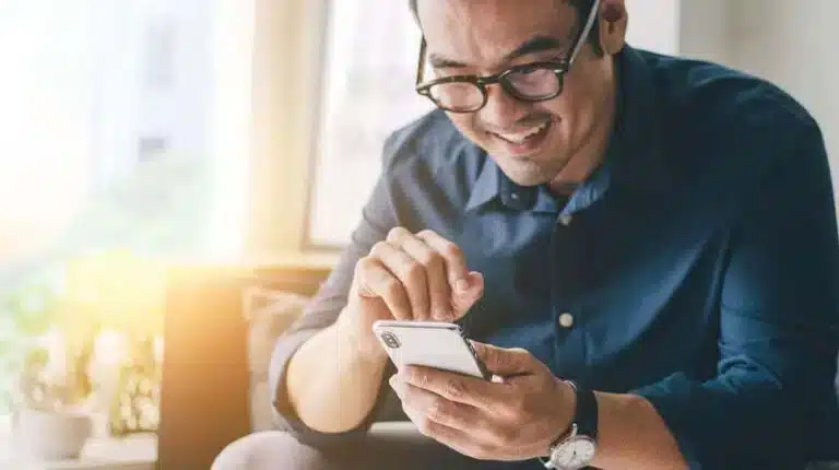 Man in glasses smiling while using a smartphone, wearing a blue shirt in a bright room.