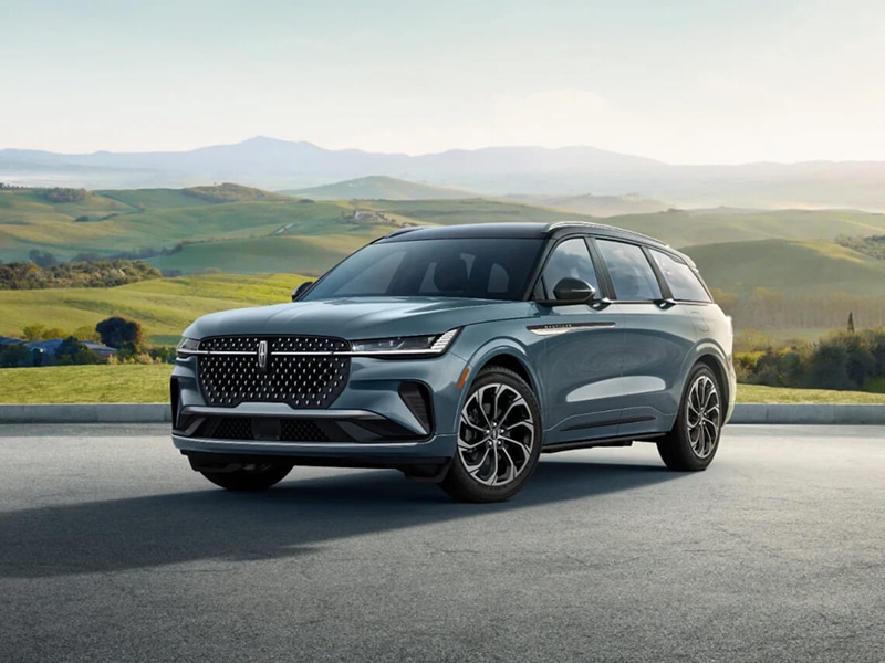 Blue SUV parked on a paved surface with rolling green hills in the background.