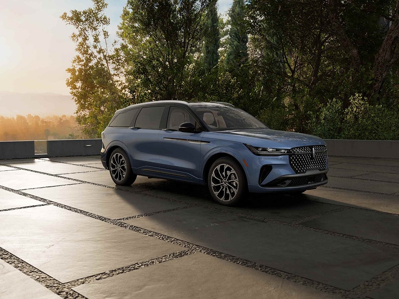 Blue SUV parked on a spacious outdoor driveway, surrounded by trees at sunset.
