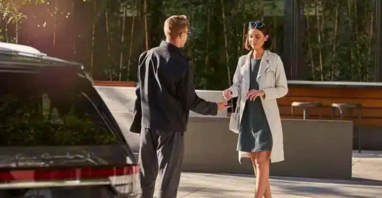 A man hands car keys to a woman in a coat outside a modern building, with a car in the foreground.