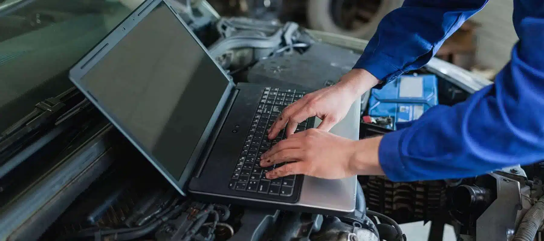 Mechanic using a laptop on a car engine, wearing a blue uniform. Car repair diagnostics.