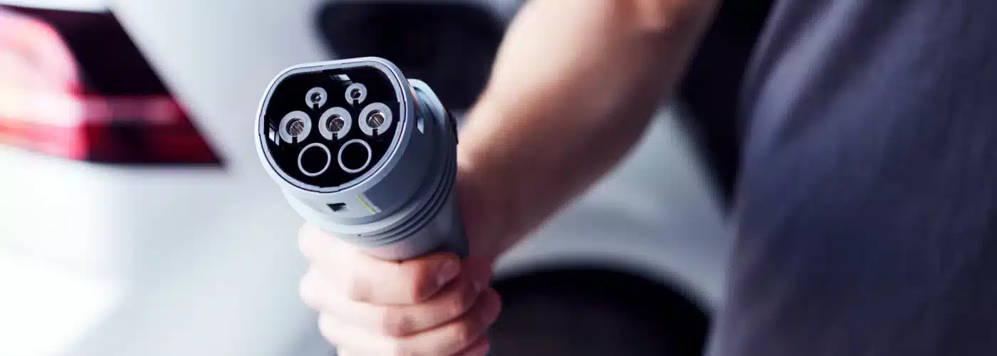 Hand holding an electric vehicle charging plug near a car, emphasizing clean energy technology.