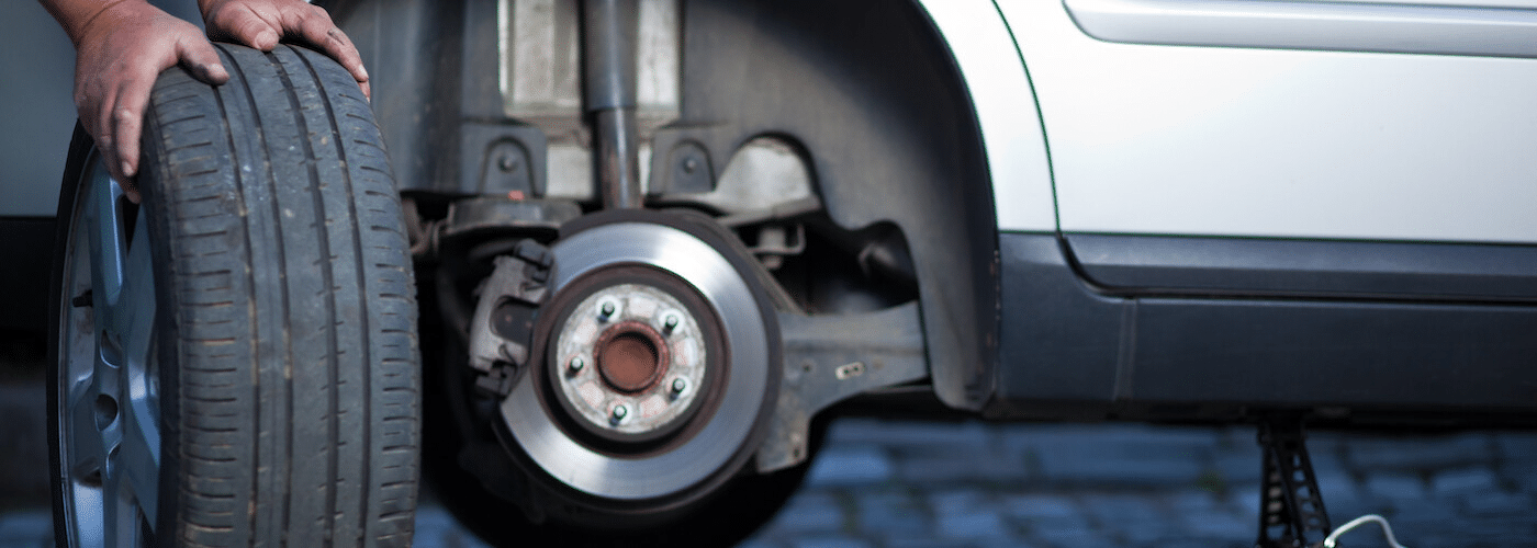 Person replacing a car tire, holding the new tire beside a raised vehicle with exposed brake disc.