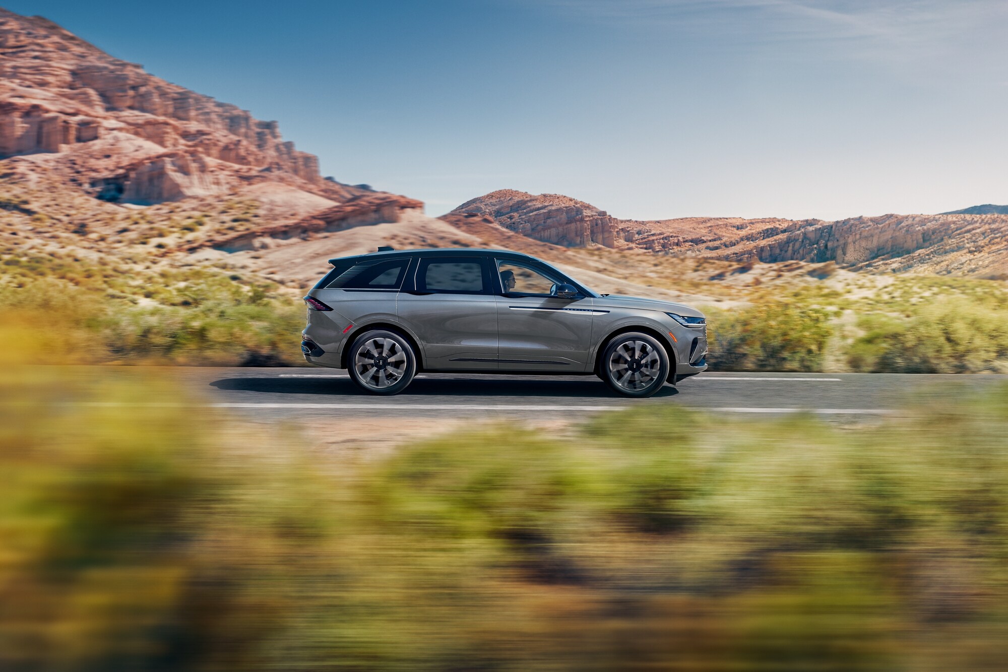 A sleek blue SUV drives on a scenic highway, surrounded by trees and a clear sky.