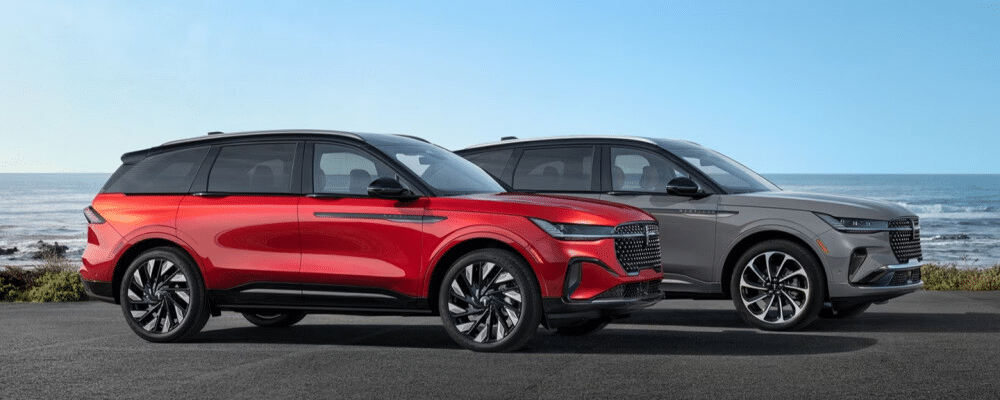 Two sleek SUVs, one red and one grey, parked by the ocean with a clear blue sky background.