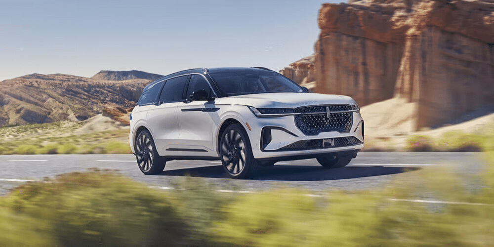 White luxury SUV driving on a desert highway with rocky hills in the background.