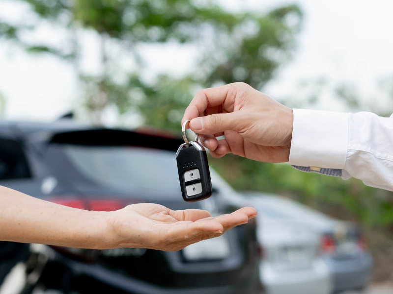 Person handing car keys to another against a blurred car background, symbolizing car sale or rental.